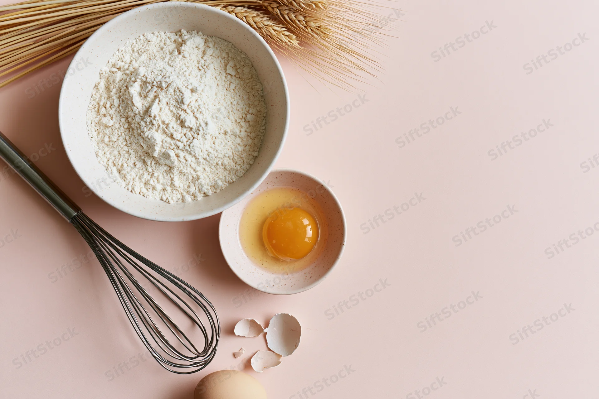 Flour and egg in bowls with whisk and wheat stalks on pink surface