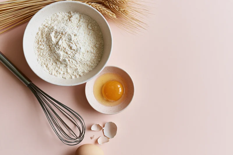 Flour and egg in bowls with whisk and wheat stalks on pink surface