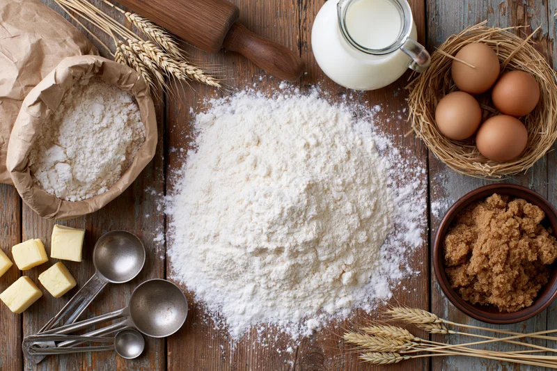 Baking ingredients arranged on a wooden table with flour, eggs, milk, sugar, butter, and wheat stalks