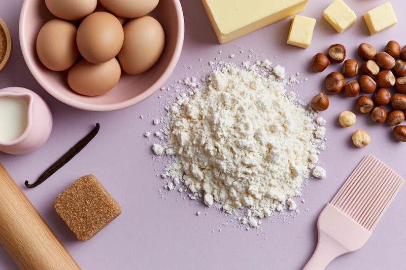 Baking ingredients arranged on a pastel surface with flour, eggs, butter, hazelnuts, sugar, vanilla, rolling pin and pastry brush