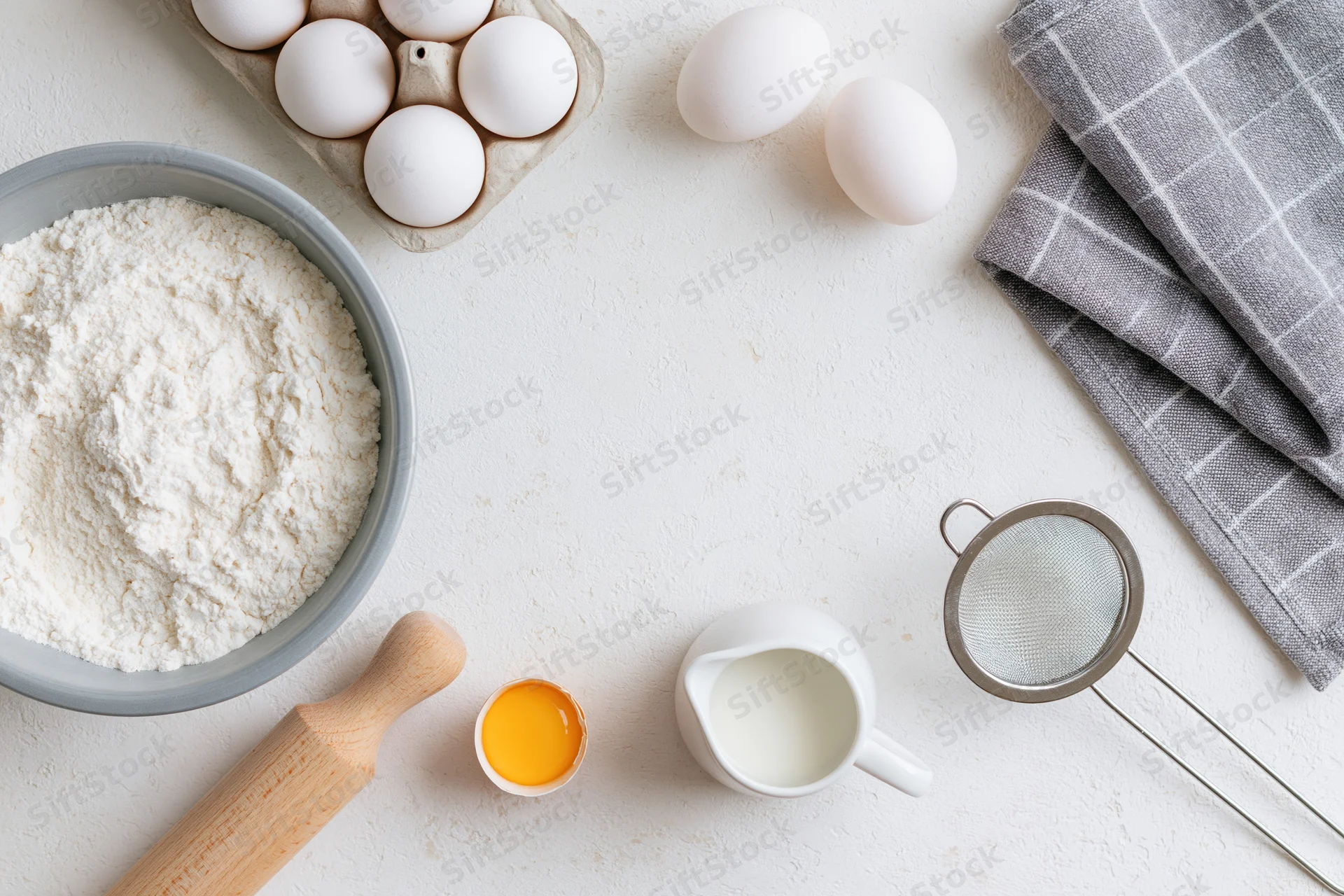 Baking ingredients arranged on a white countertop with rolling pin, sieve, and checkered napkin