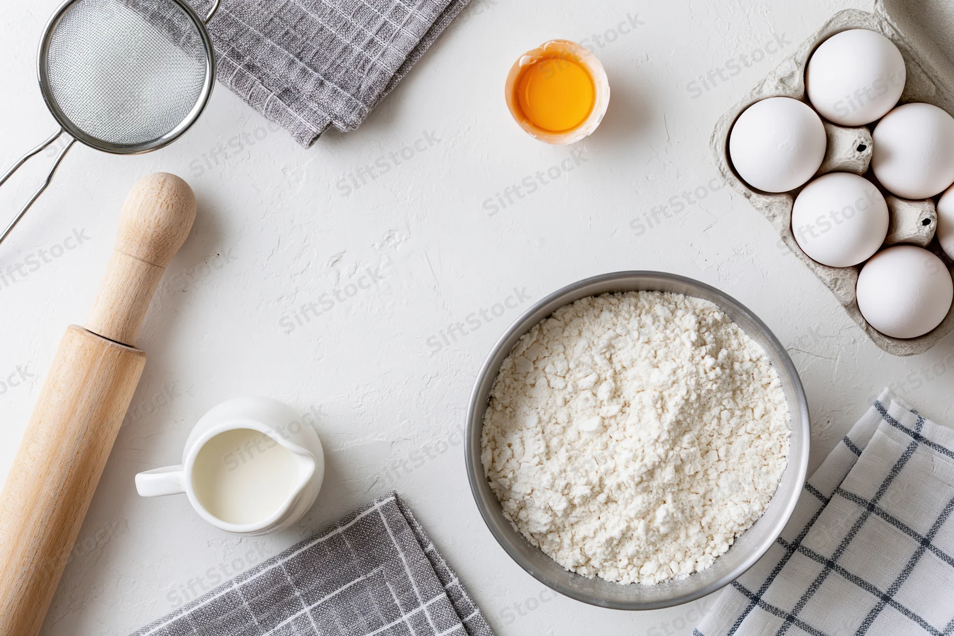 Baking ingredients arranged on a white countertop with rolling pin, bowl of flour, eggs, milk and sieve