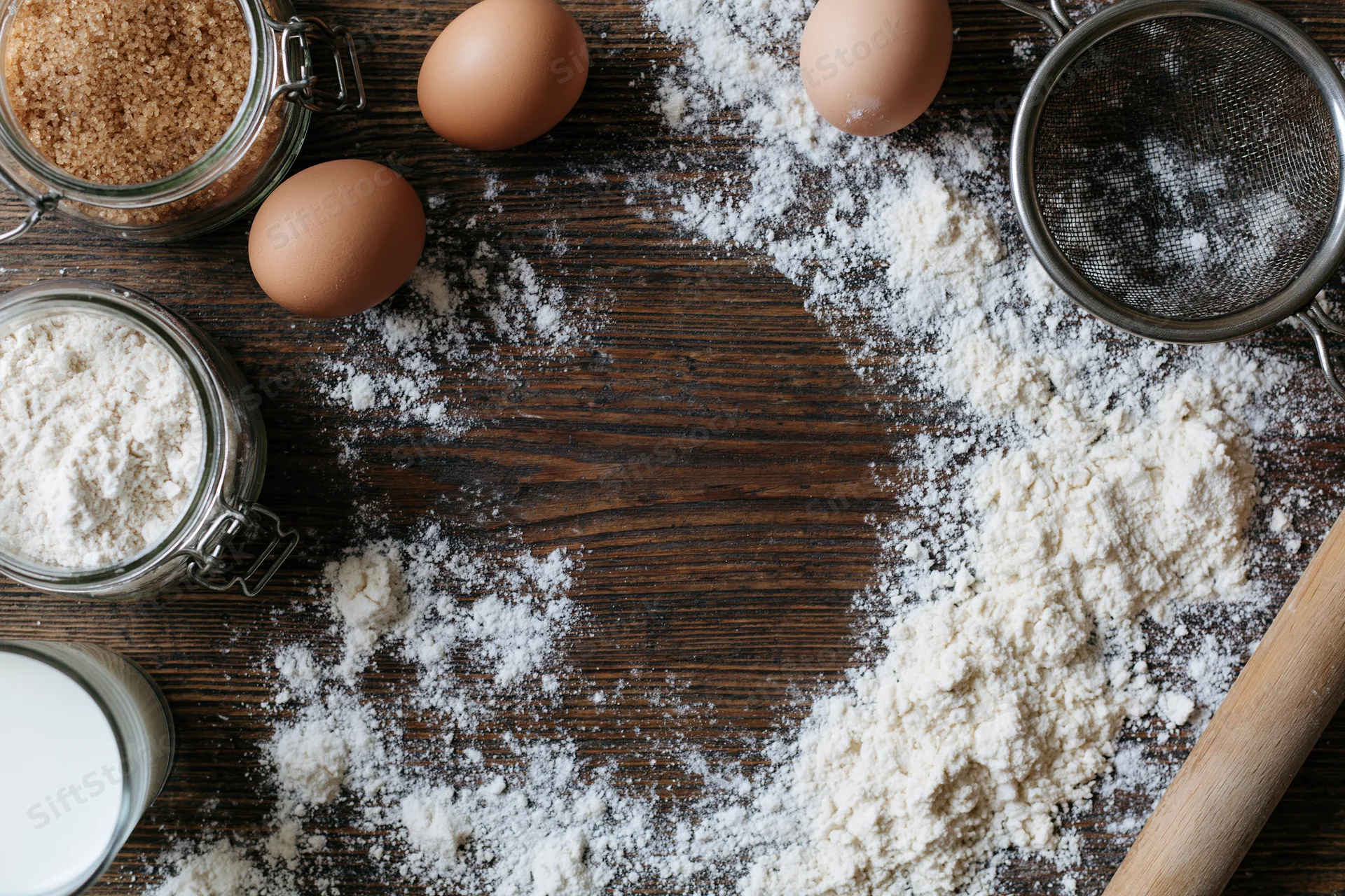 Baking ingredients scattered on a wooden countertop with flour, eggs, sugar, milk, and a rolling pin