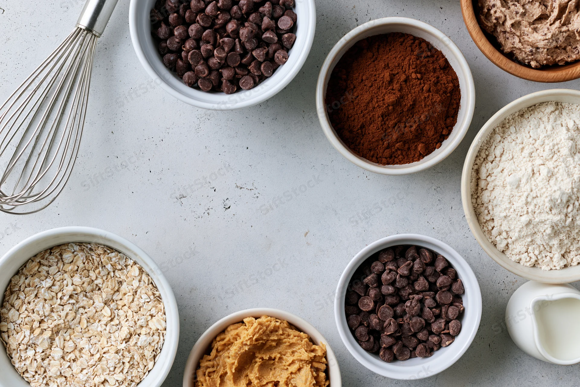 Baking ingredients arranged in white bowls on a light countertop with a whisk and milk pitcher, overhead view