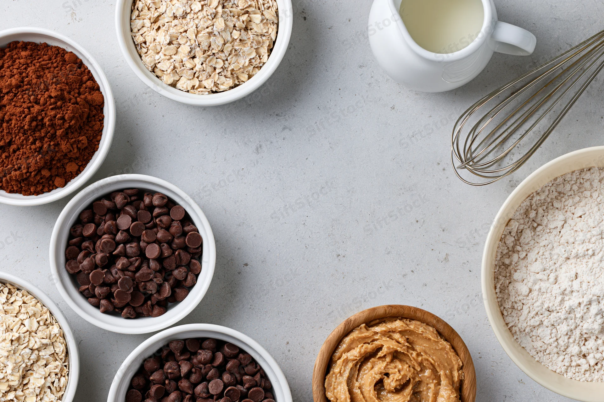 Various baking ingredients arranged in bowls on a light gray countertop with a whisk and milk jug nearby