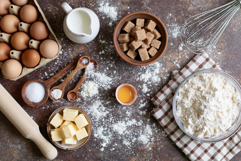 Baking ingredients arranged on a rustic countertop with flour dusted around and a checkered cloth under a bowl of flour
