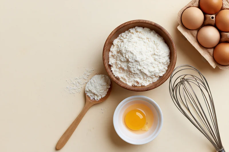 Baking ingredients arranged on a beige surface with flour in a wooden bowl, eggs in a carton, a whisk, and a spoon with flour, viewed from above