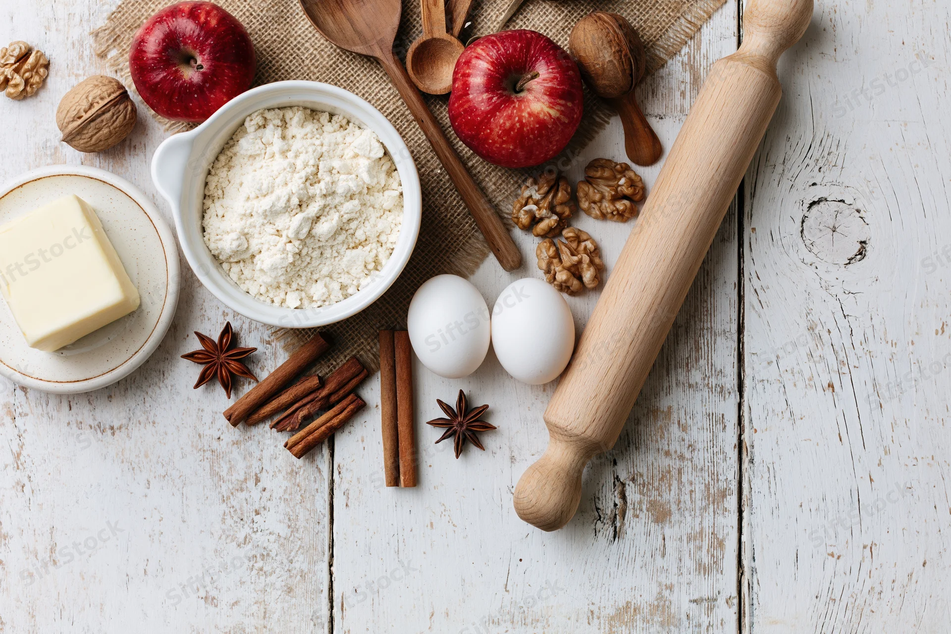 Baking ingredients arranged on a rustic white wooden table with apples, flour, eggs, butter, walnuts, cinnamon, and a rolling pin