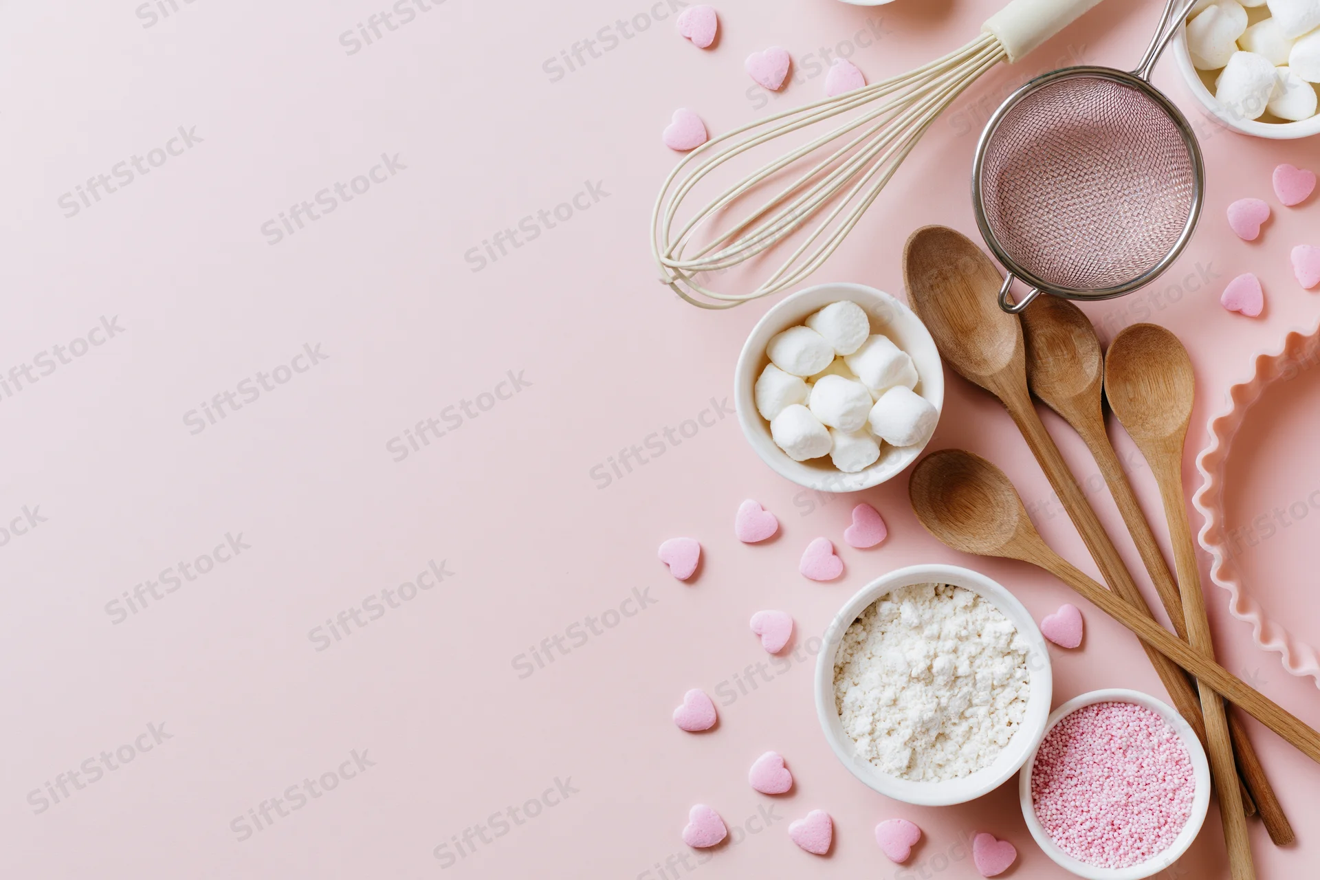 Baking ingredients and utensils arranged on a pink surface with heart-shaped candies and copyspace