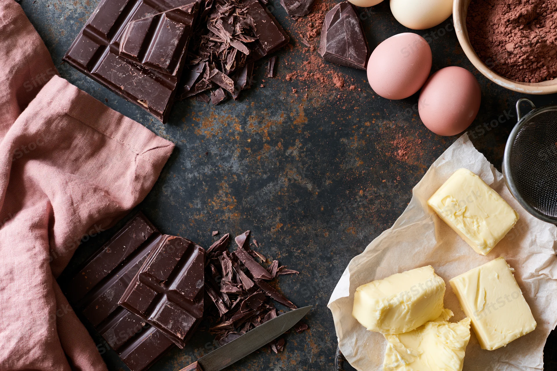 Chocolate bars and chunks, butter, eggs, cocoa powder arranged on dark textured surface with knife and parchment paper for baking preparation