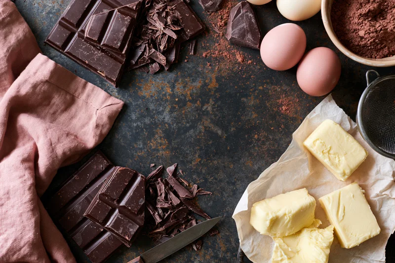 Chocolate bars and chunks, butter, eggs, cocoa powder arranged on dark textured surface with knife and parchment paper for baking preparation