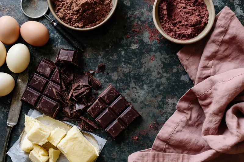 Chocolate bars and baking ingredients arranged on a dark rustic countertop with butter, eggs, cocoa powder, and a pink cloth