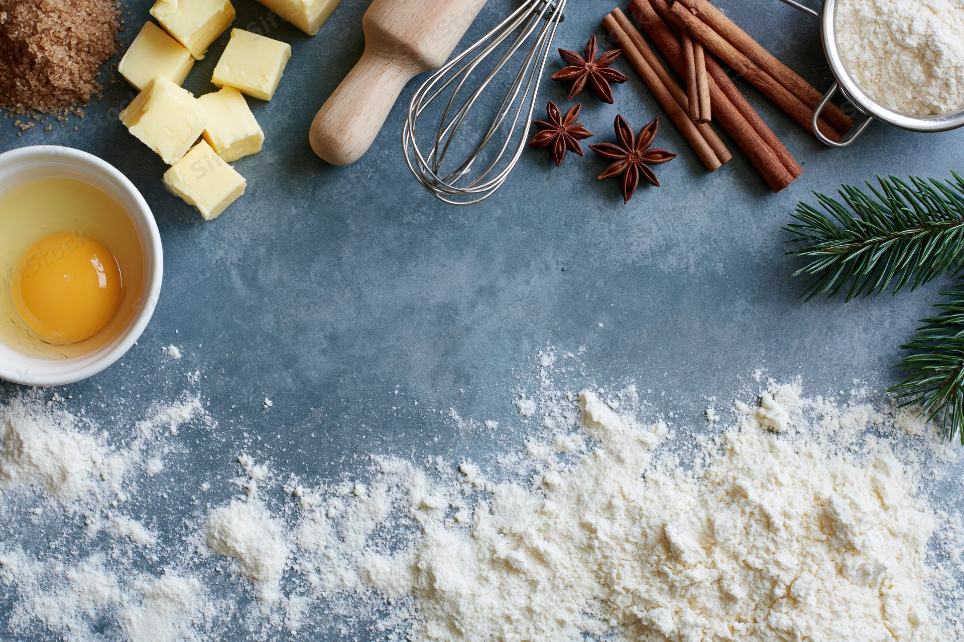 Baking ingredients arranged on a gray countertop with flour, egg, butter, cinnamon, star anise, and pine sprigs for christmas holiday dessert preparation