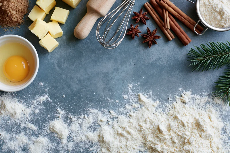 Baking ingredients arranged on a gray countertop with flour, egg, butter, cinnamon, star anise, and pine sprigs for christmas holiday dessert preparation