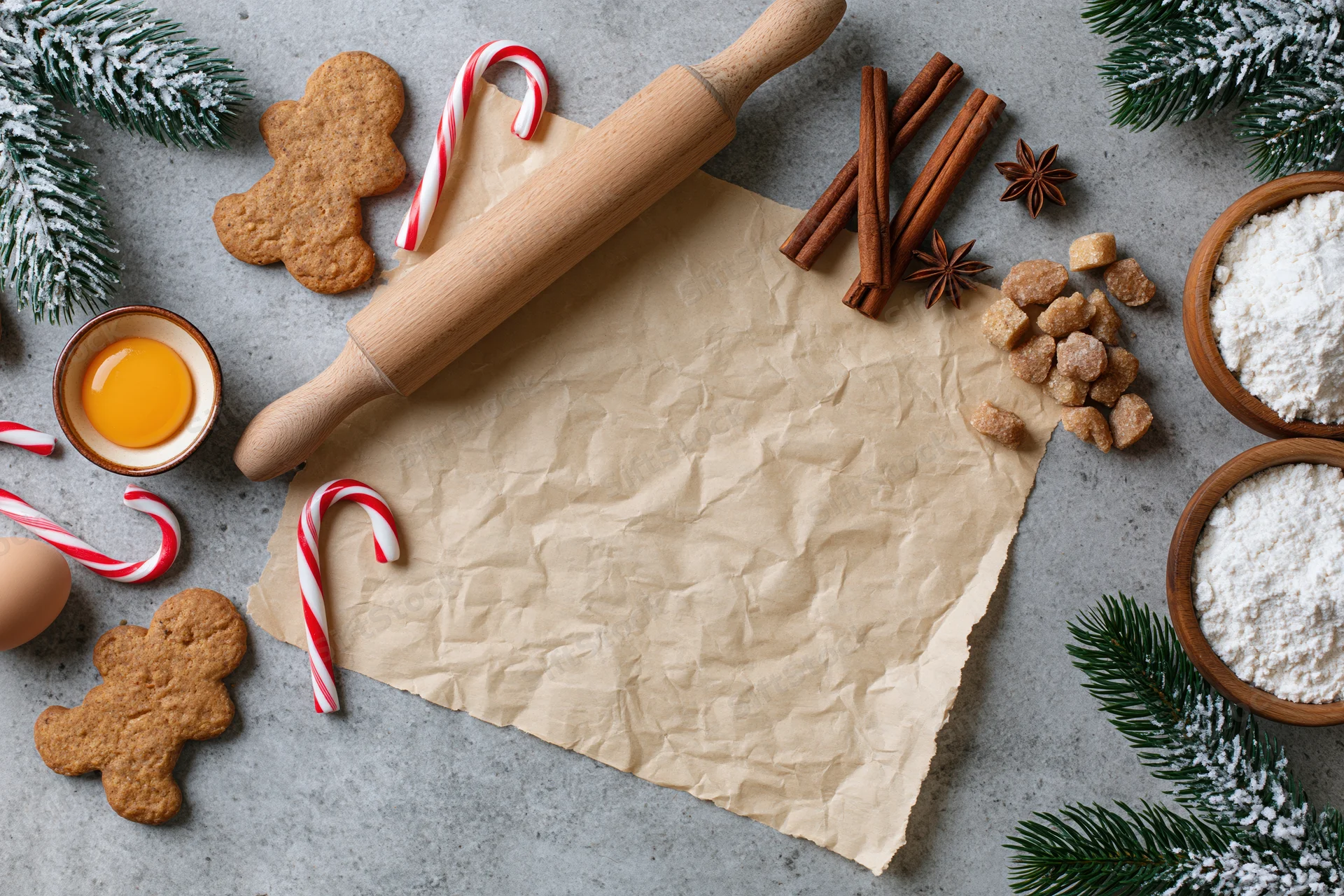 Gingerbread cookies and baking ingredients arranged on a countertop with pine branches and candy canes for christmas