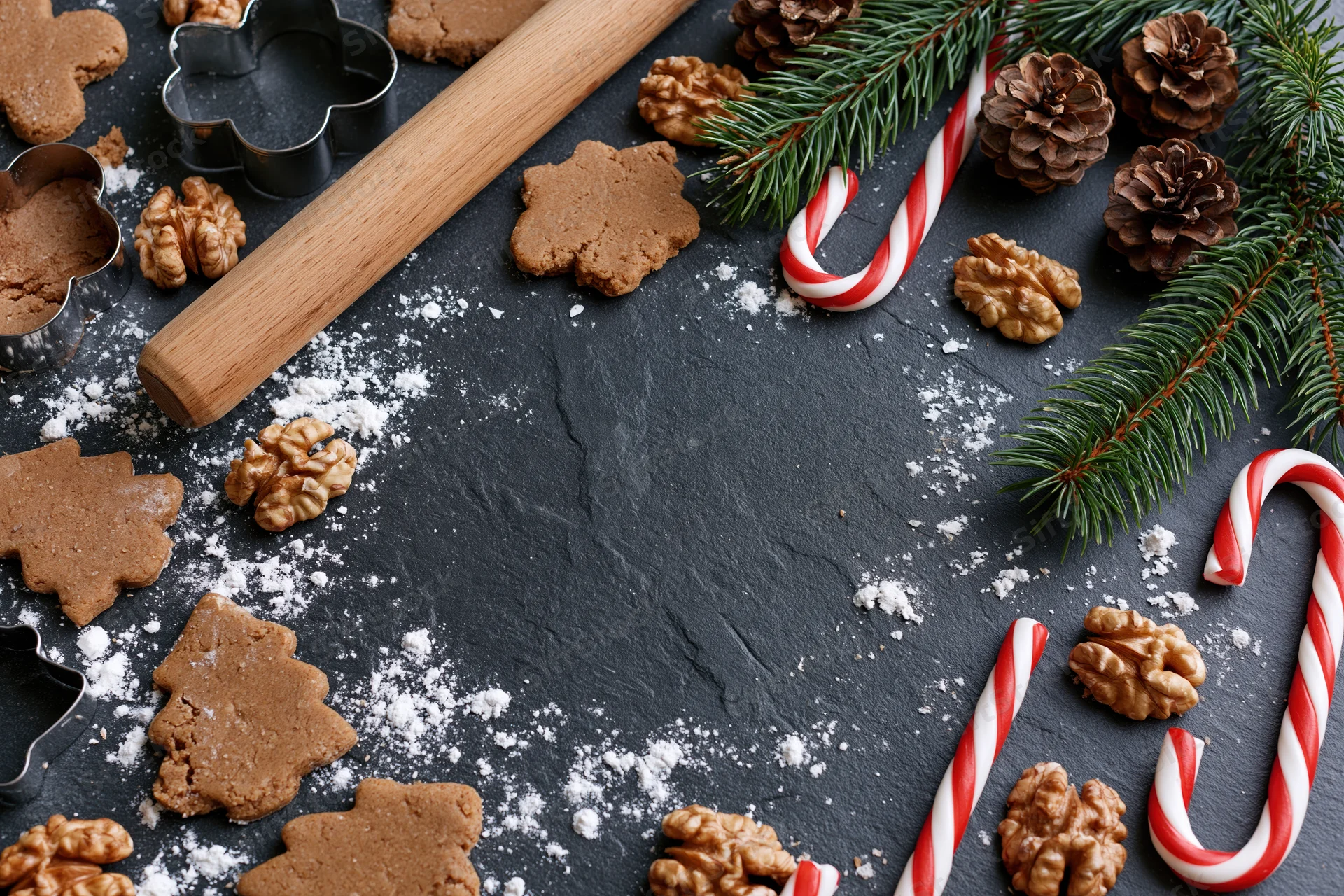 Gingerbread cookies shaped like trees and stars with candy canes, pine cones, walnuts and cookie cutters on a dark slate surface dusted with flour