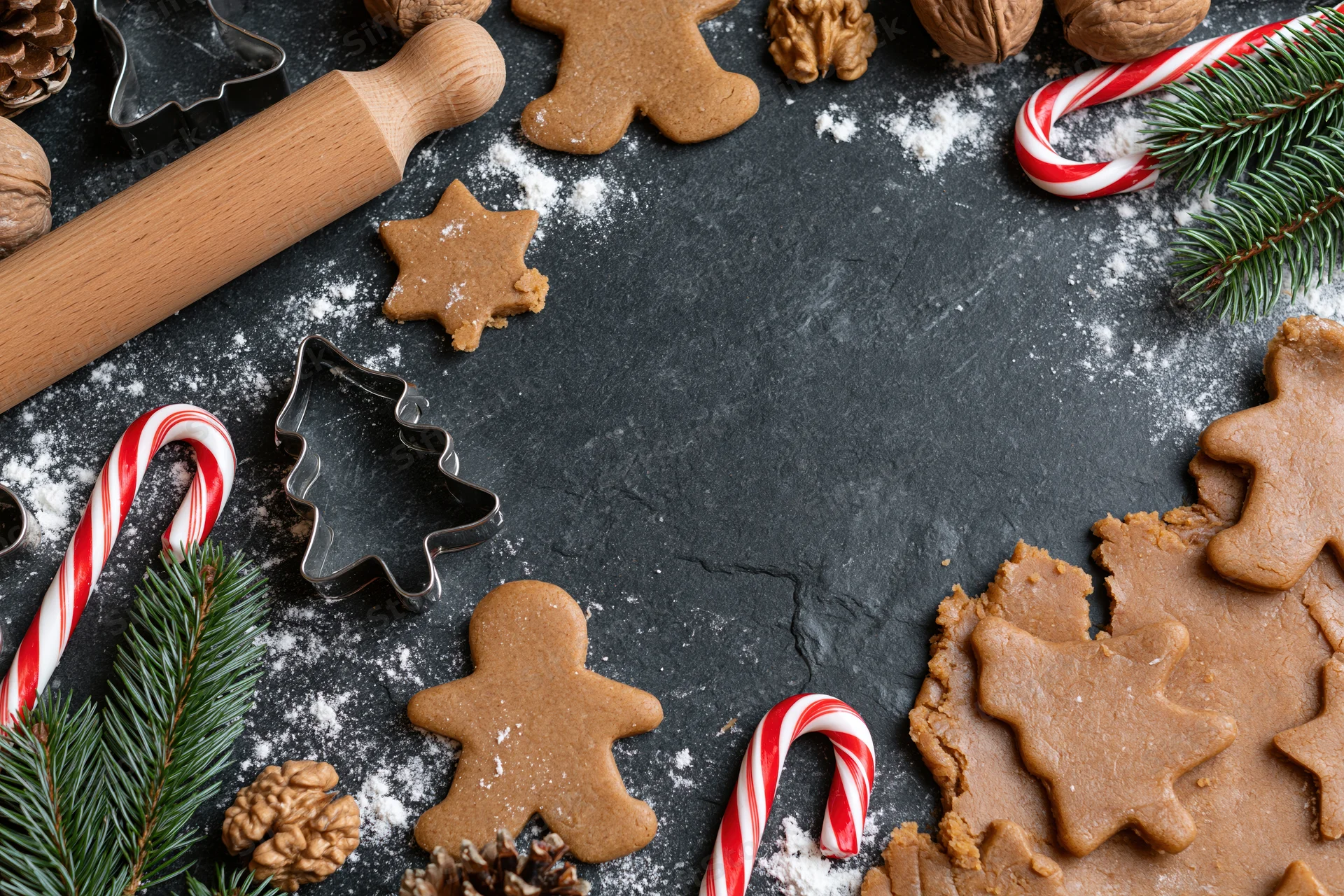 Gingerbread cookies and baking tools arranged on dark surface with candy canes and pine branches