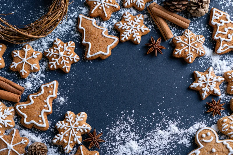 Gingerbread cookies shaped as snowflakes and trees arranged around a dark blue surface with cinnamon sticks, pinecones, and powdered sugar, leaving copyspace in the center
