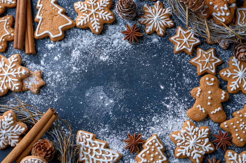 Gingerbread cookies shaped as snowflakes, trees, and gingerbread men arranged around a dark surface dusted with flour, with cinnamon sticks and pinecones, creating a festive holiday border with central copyspace