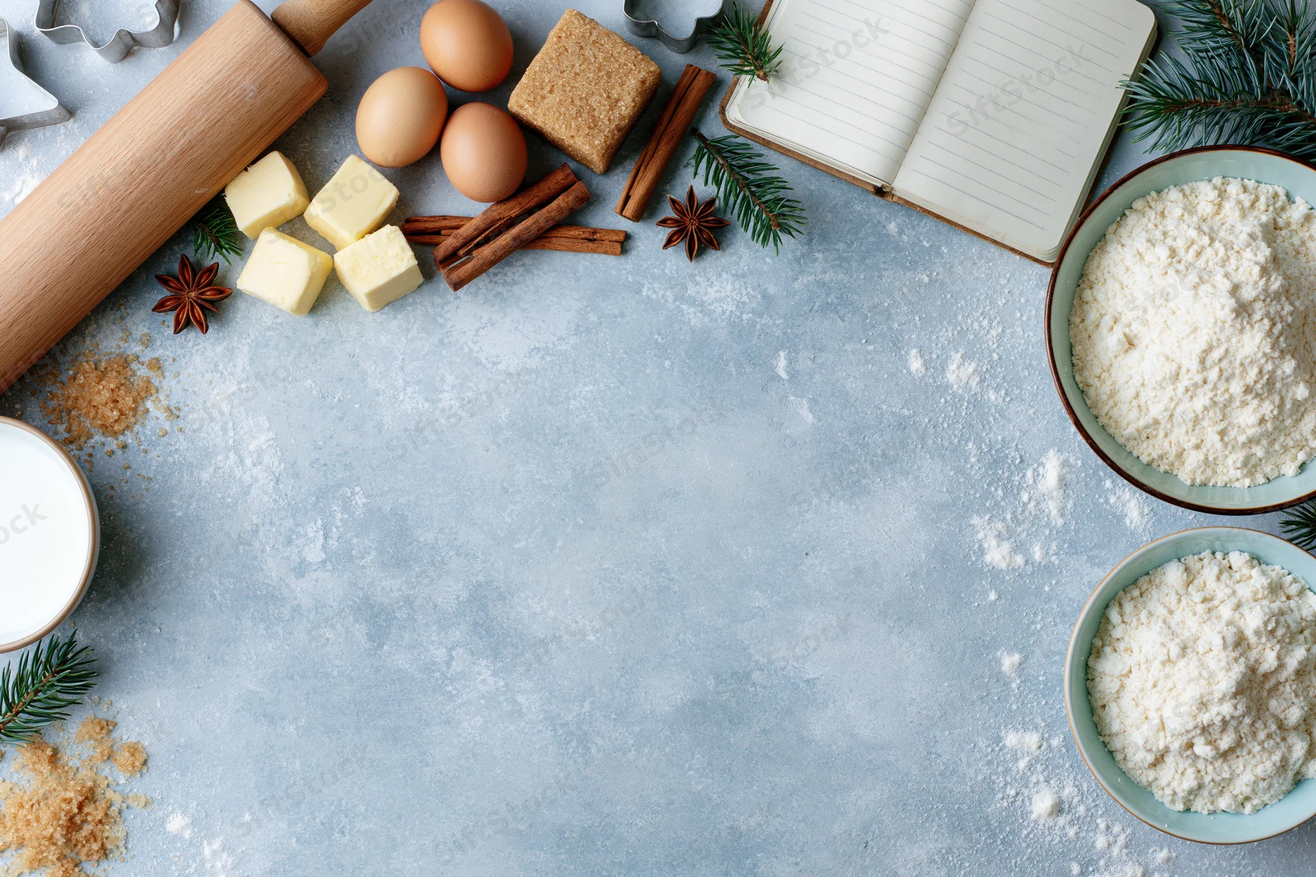 Baking ingredients arranged on a floured countertop with evergreen sprigs and a notebook for holiday recipe planning