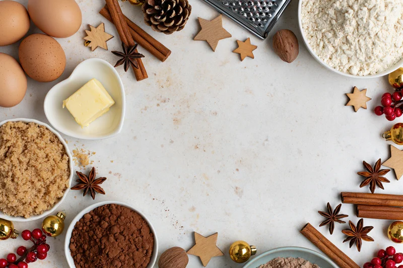 Baking ingredients arranged around a white surface with christmas decorations and copyspace in center