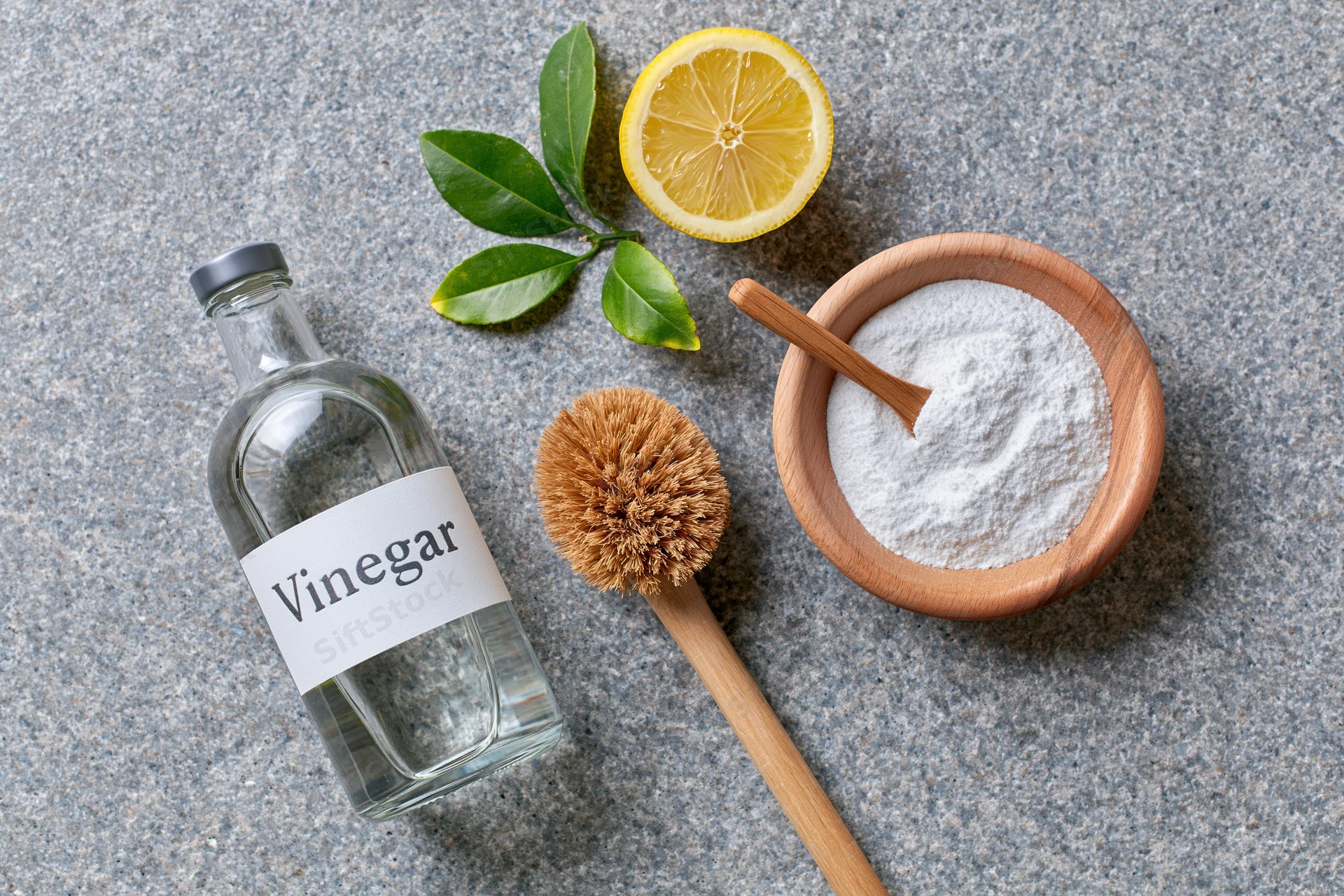 Vinegar bottle, lemon slice, baking soda in wooden bowl, and coconut fiber brush arranged on gray stone countertop