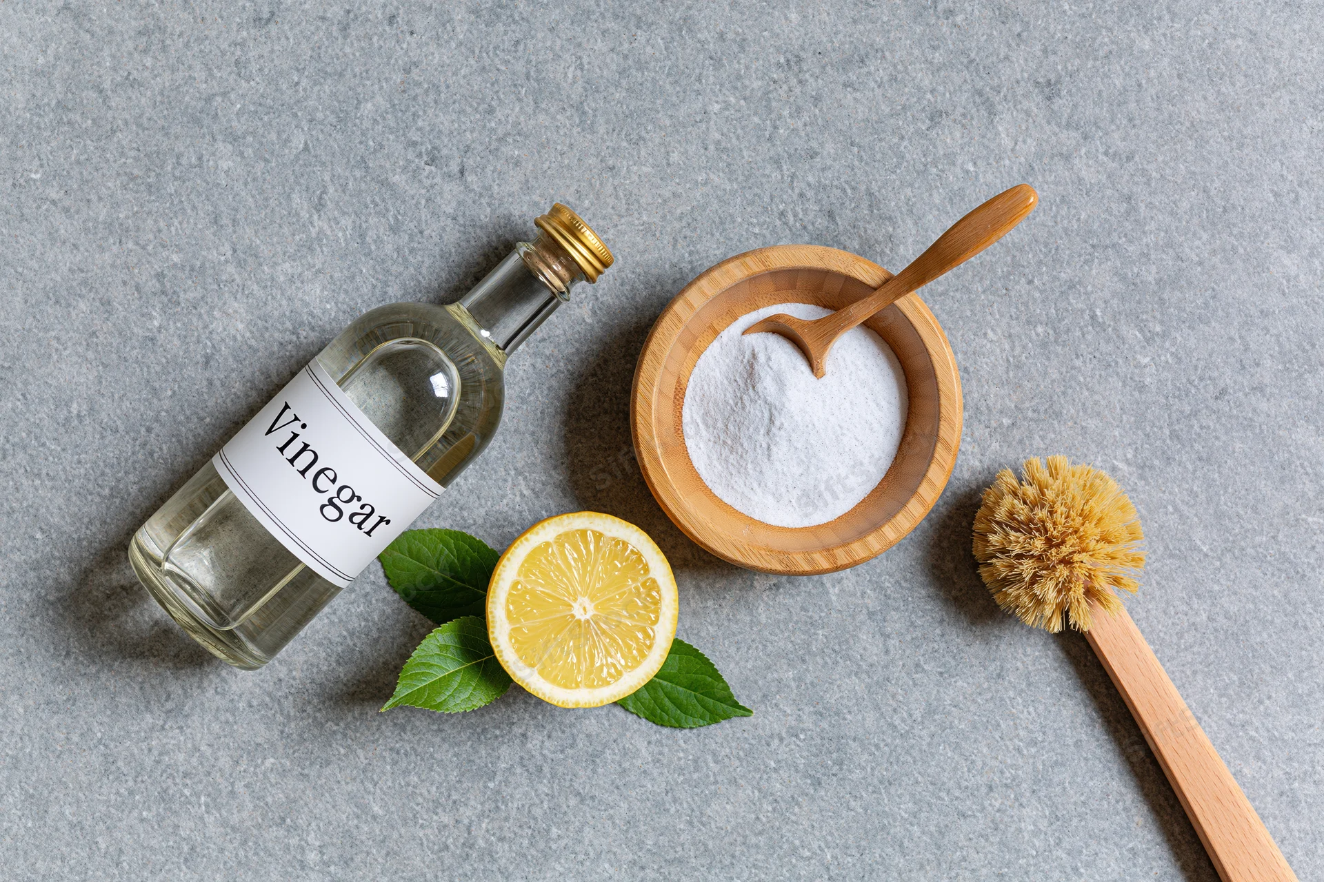 Vinegar bottle, lemon slice, baking soda in wooden bowl, and cleaning brush arranged on gray surface