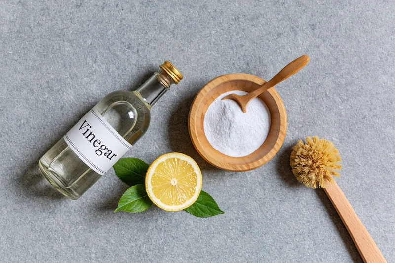 Vinegar bottle, lemon slice, baking soda in wooden bowl, and cleaning brush arranged on gray surface