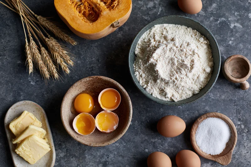 Baking ingredients arranged on dark countertop including flour, eggs, butter, pumpkin, wheat, and salt