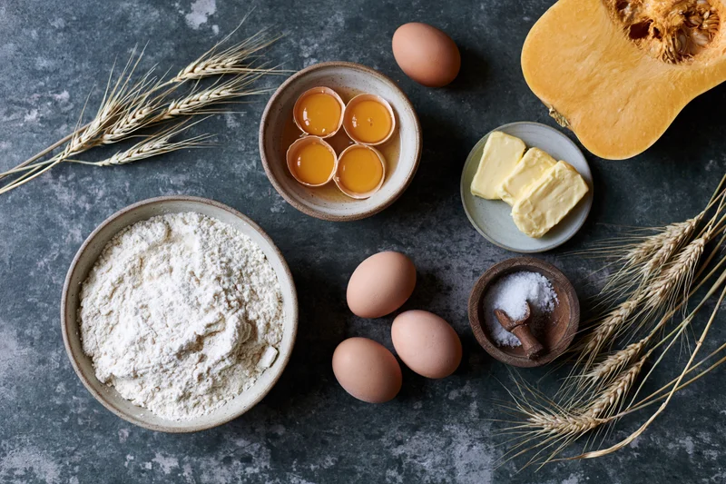Baking ingredients arranged on dark stone surface with wheat stalks and pumpkin