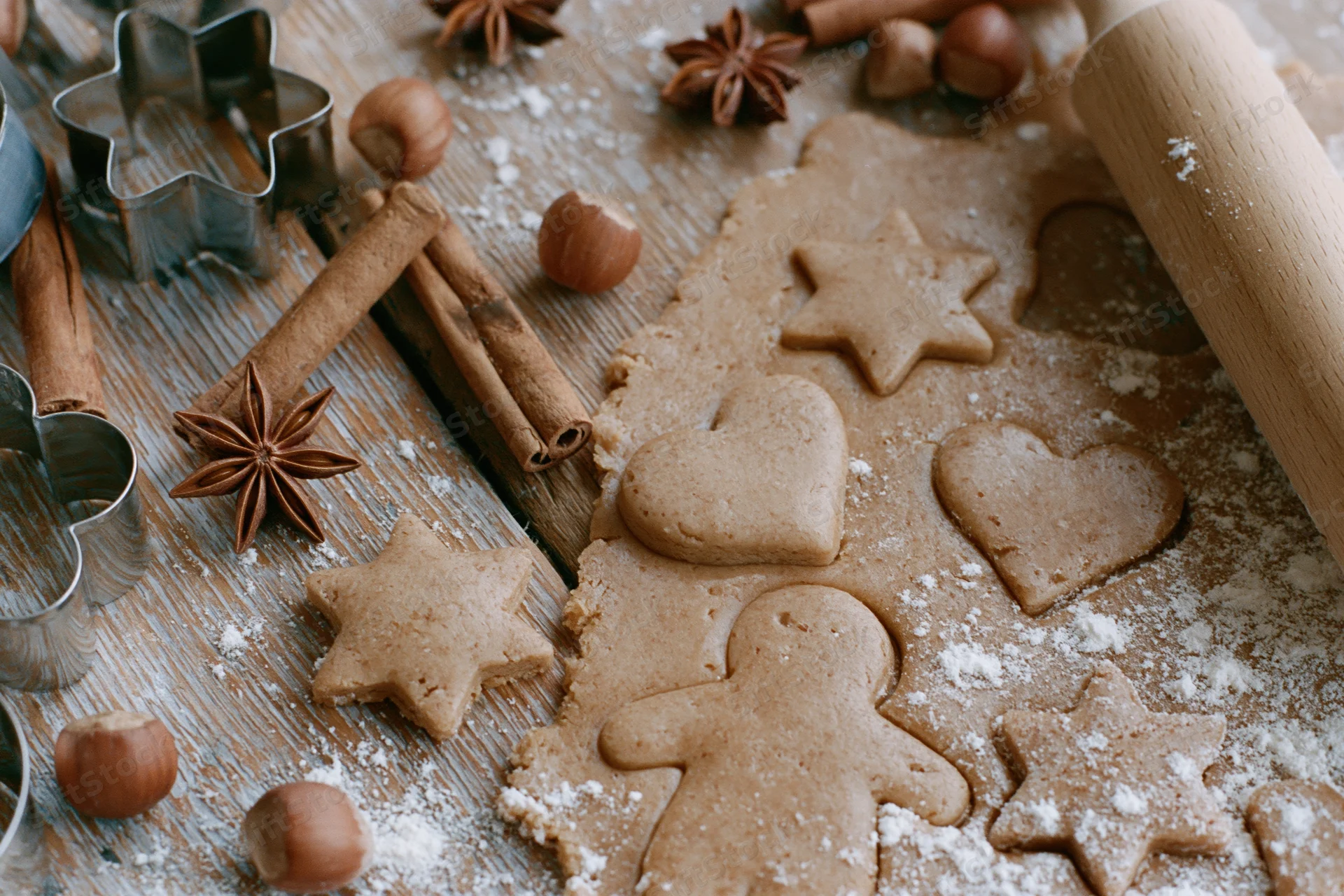 Gingerbread dough cut into star, heart and gingerbread man shapes on a floured wooden table with cinnamon sticks, hazelnuts and cookie cutters around