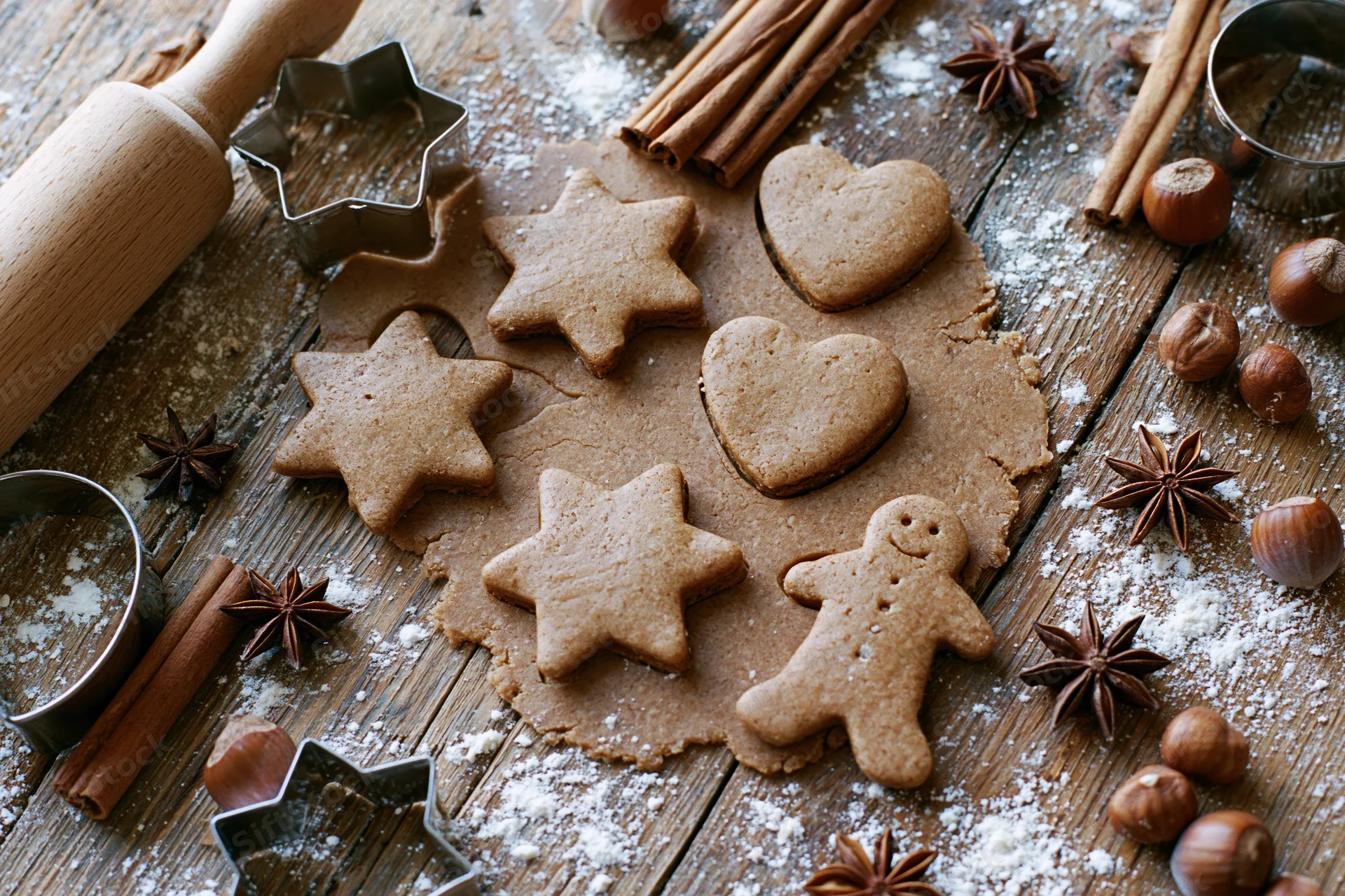 Gingerbread cookies shaped as stars, hearts and a gingerbread man on floured wooden surface with spices and baking tools around