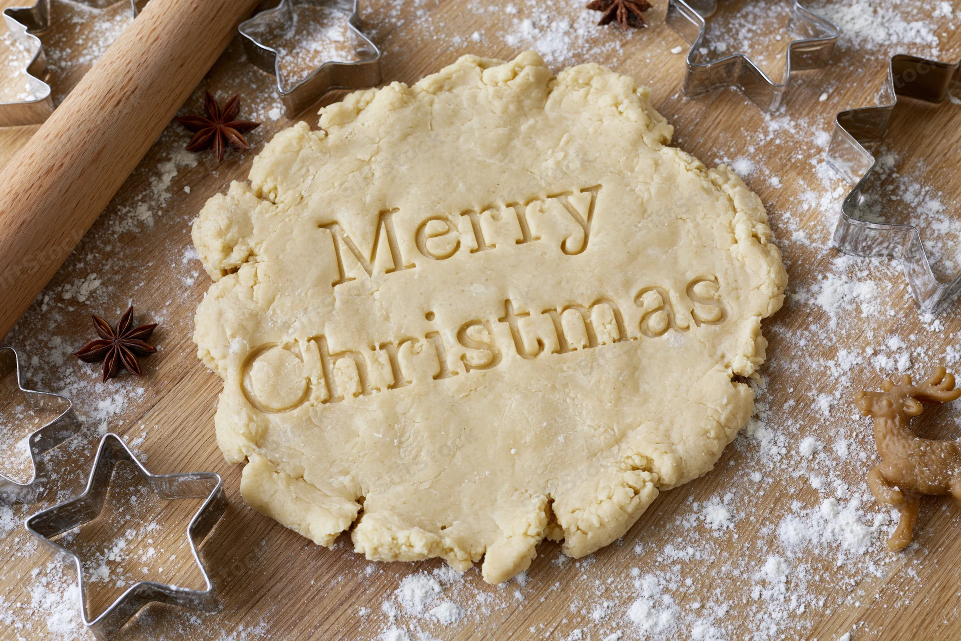 Cookie dough with "Merry Christmas" imprinted on it surrounded by star-shaped cutters and anise stars on a floured wooden surface