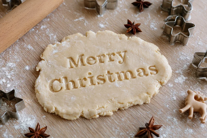 Flattened cookie dough with "Merry Christmas" imprinted on wooden table surrounded by star-shaped cutters and spices
