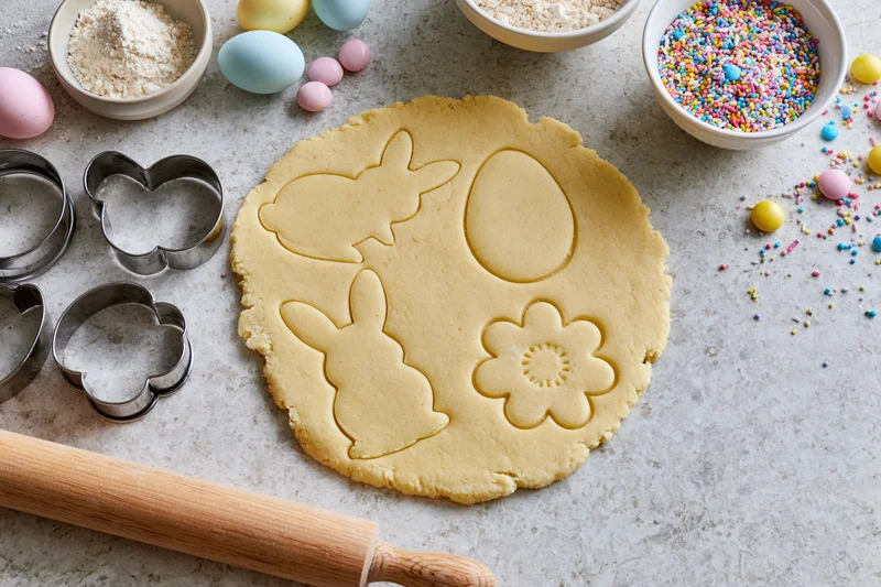 Easter cookie dough with bunny and flower shapes cut out on a countertop surrounded by baking tools and colorful eggs