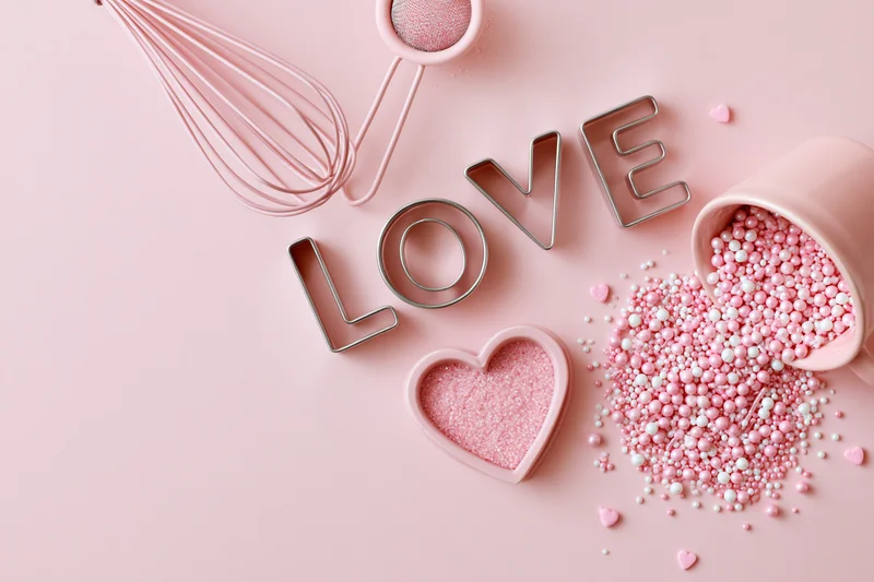 Pink baking tools and sprinkles arranged with love cookie cutters on a pastel surface