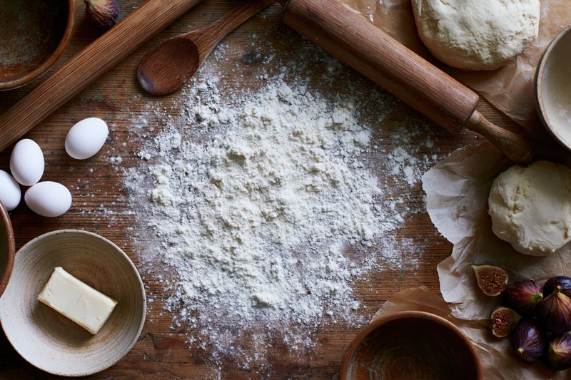 Flour scattered on a wooden countertop with baking ingredients arranged around it
