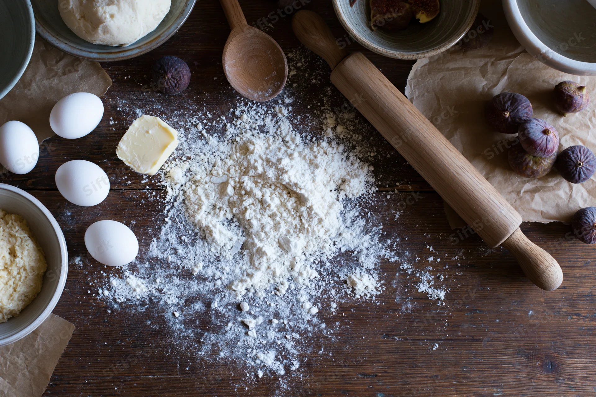 Flour and baking ingredients arranged on a wooden table with rolling pin, eggs, butter, and figs
