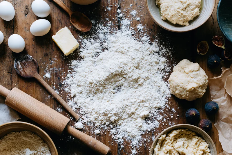 Flour and baking ingredients arranged on a wooden table with dough, eggs, butter, and figs