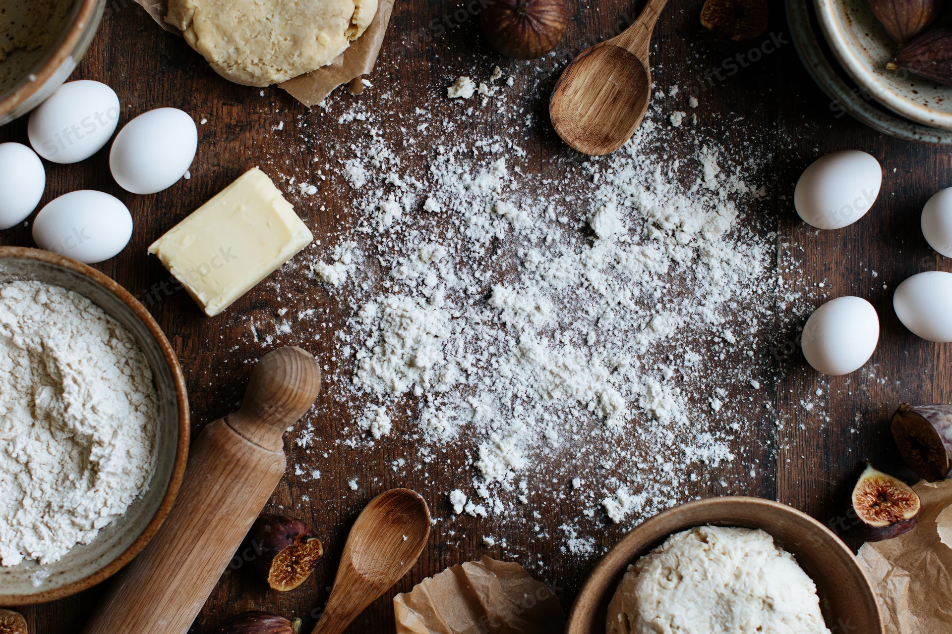 Baking ingredients arranged on a wooden table with flour, eggs, butter, rolling pin and figs in a flat lay overhead view