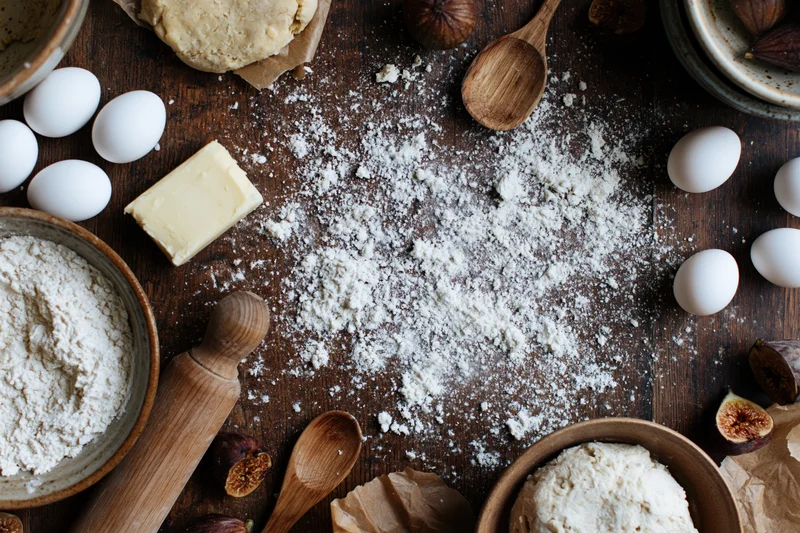 Baking ingredients arranged on a wooden table with flour, eggs, butter, rolling pin and figs in a flat lay overhead view