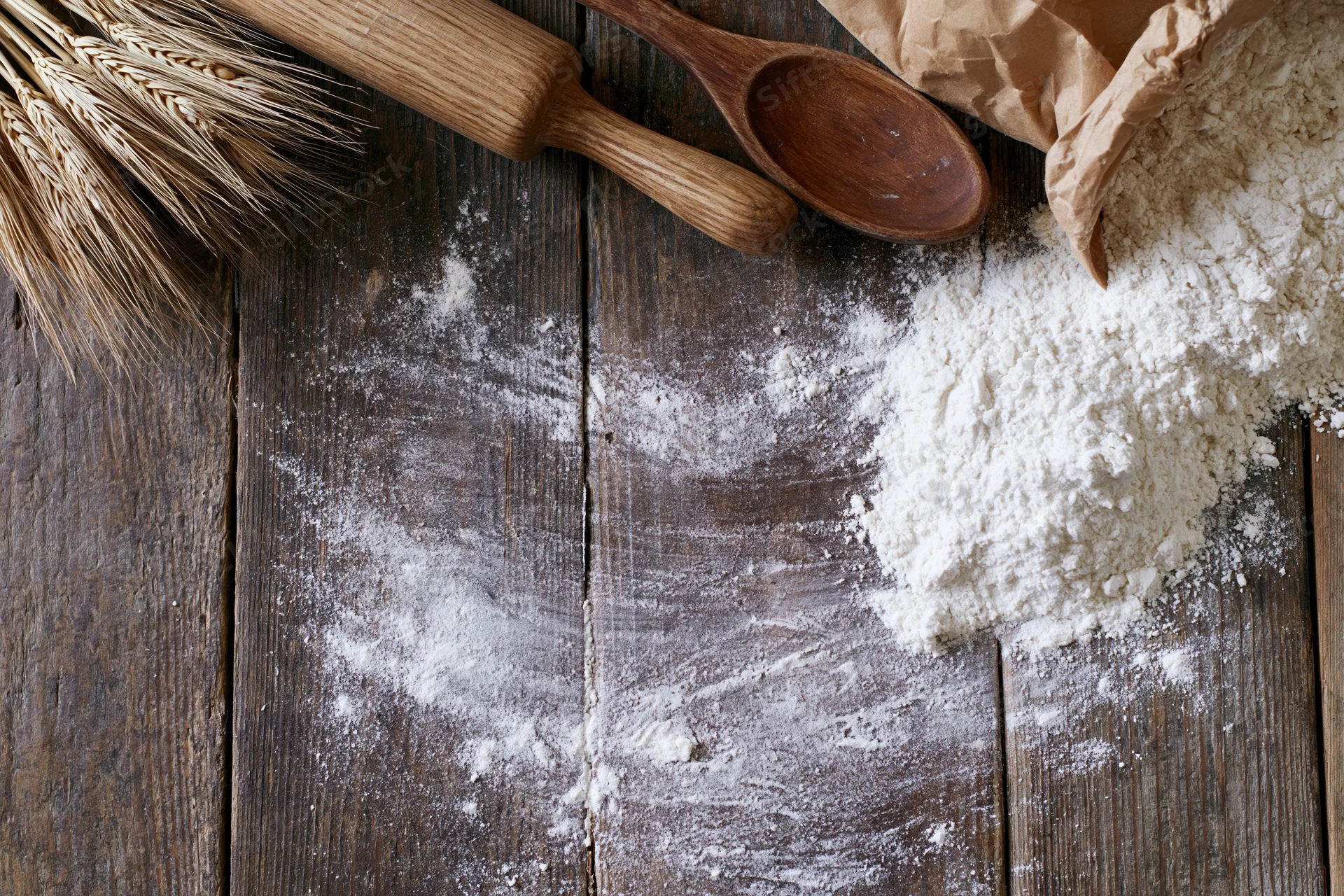 Flour spilled on a wooden table beside wheat stalks, a rolling pin and a wooden spoon
