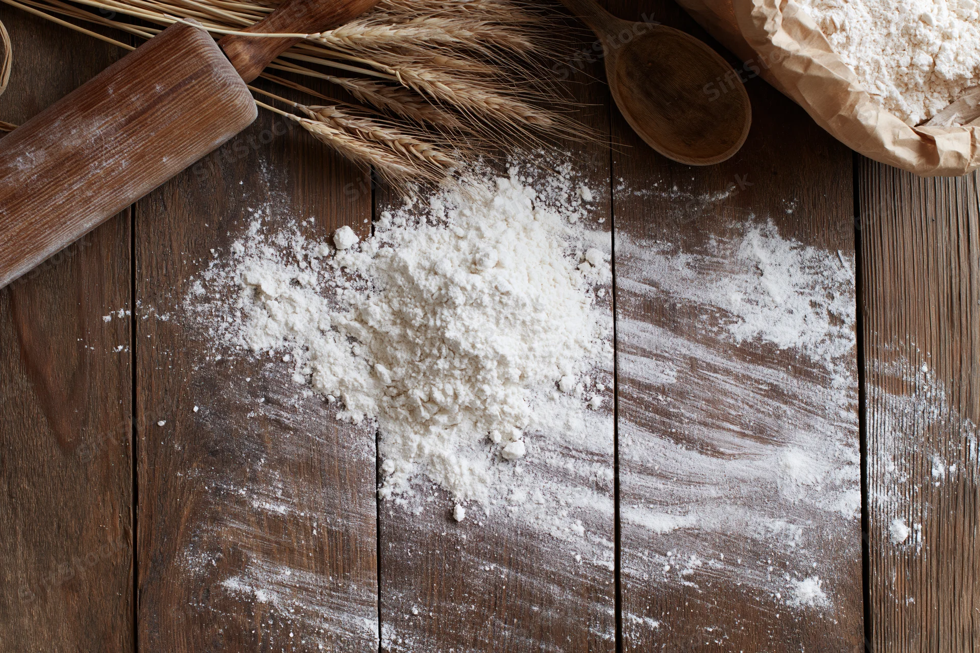 Flour scattered on a wooden table with wheat stalks, rolling pin and spoon nearby