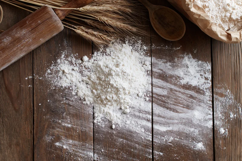 Flour scattered on a wooden table with wheat stalks, rolling pin and spoon nearby