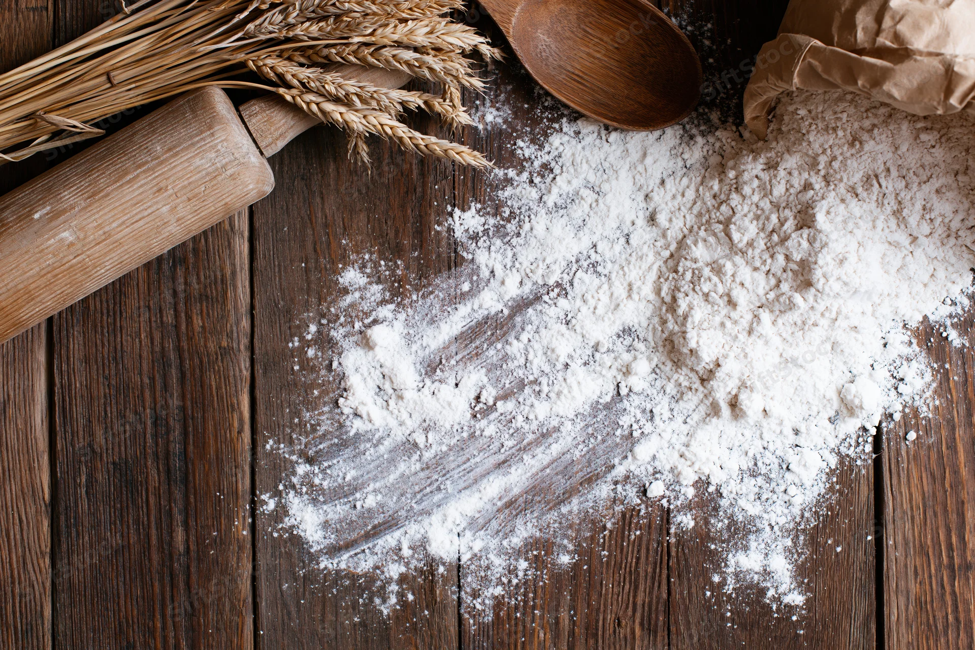 Flour spilled on a wooden table beside a rolling pin, wheat stalks, and a wooden spoon