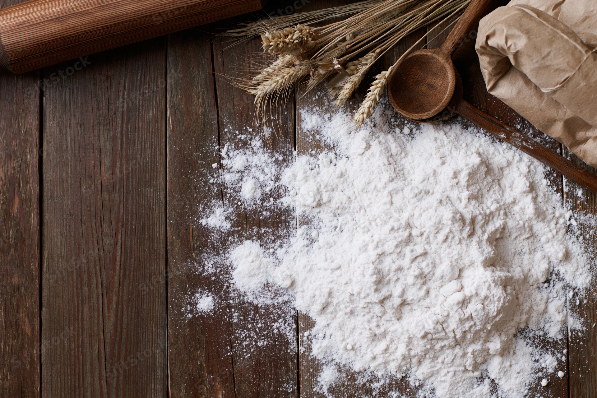 Pile of white flour on dark wooden table with wheat stalks, wooden spoon, rolling pin and paper bag nearby