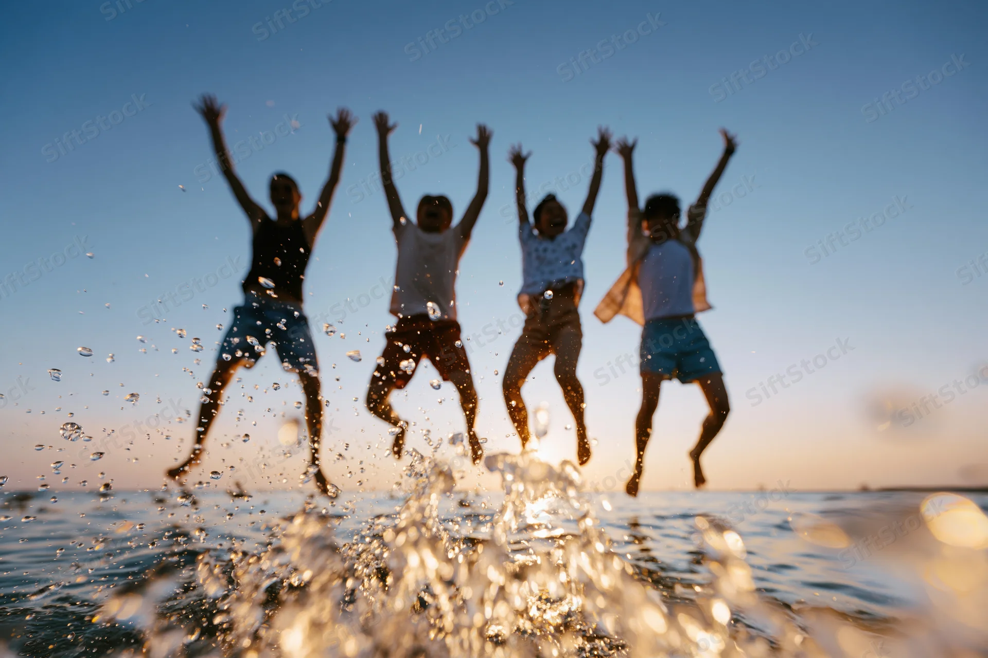 Four friends jumping in unison at the beach with water splashing in the foreground at sunset