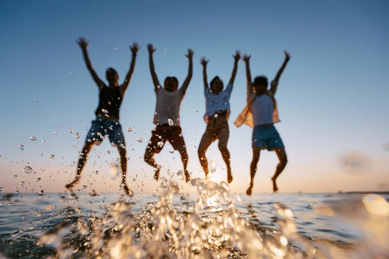 Four friends jumping in unison at the beach with water splashing in the foreground at sunset