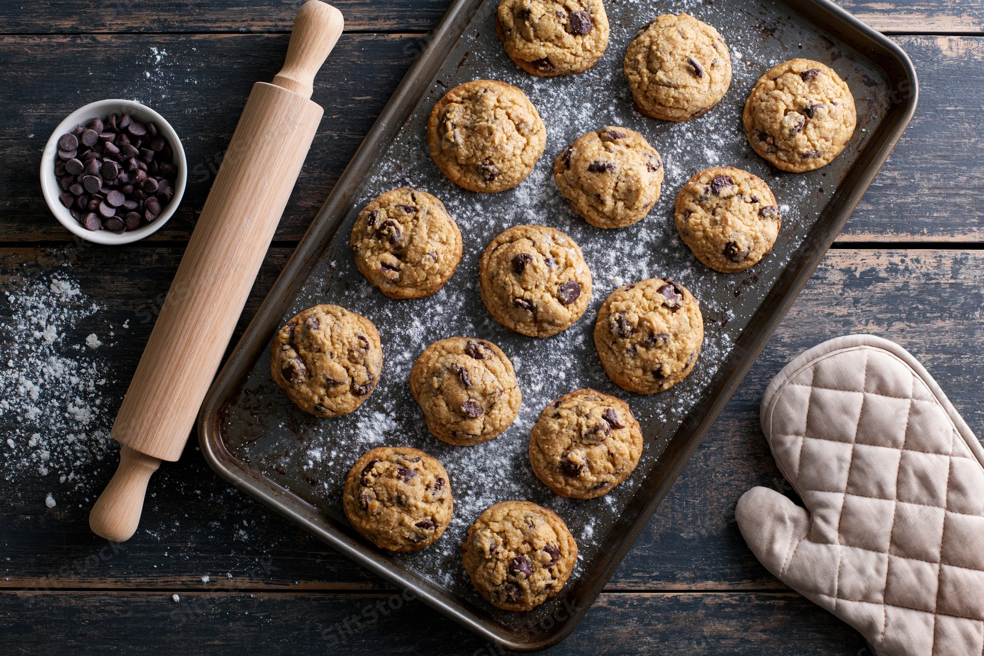 Freshly baked chocolate chip cookies on a baking tray with rolling pin and oven mitt on dark wooden surface
