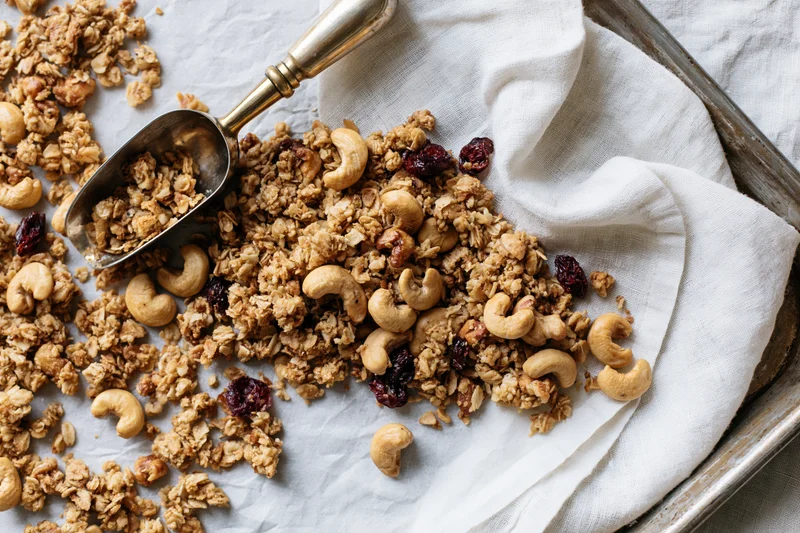 Granola with cashews and dried cranberries on a parchment-lined baking tray with a metal scoop and linen cloth